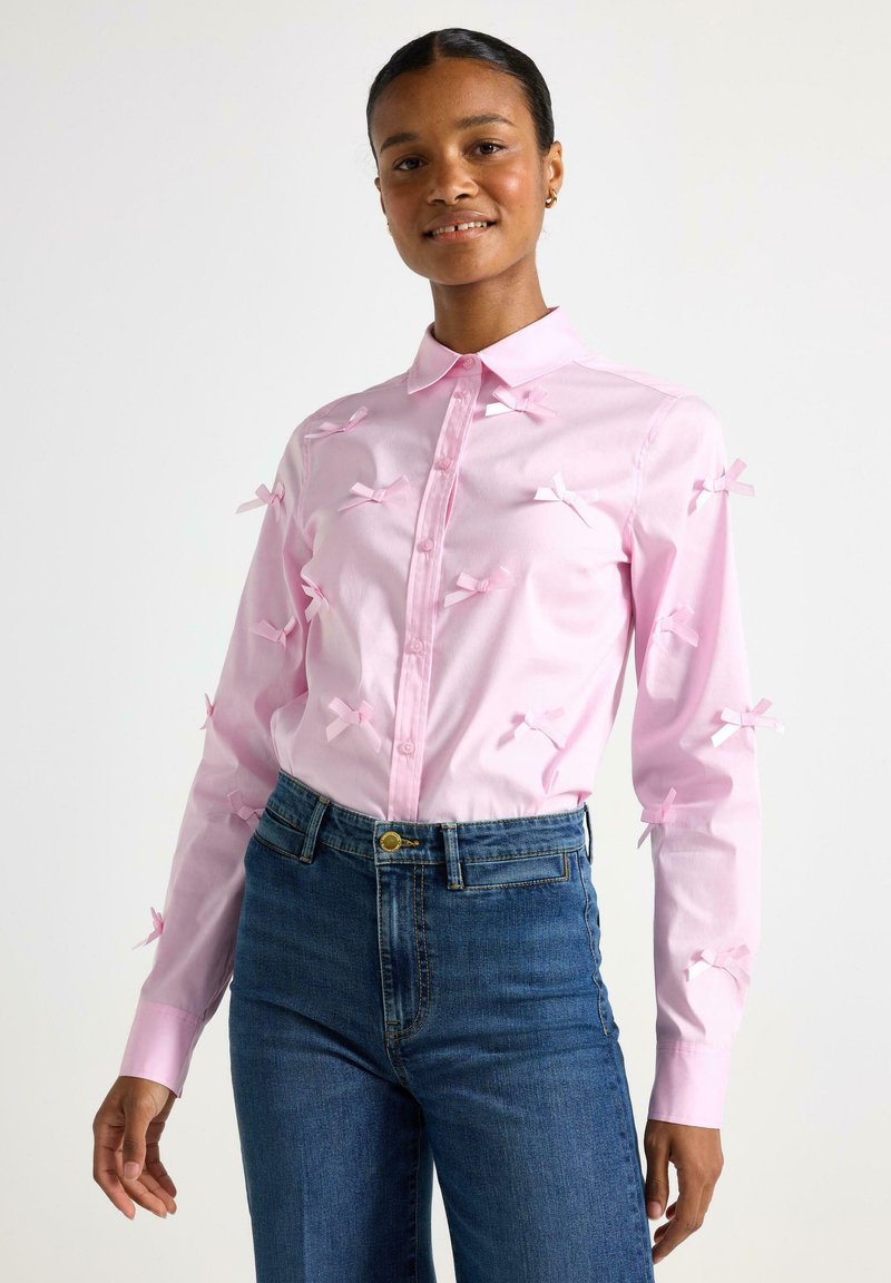 Camicia rosa con bottoni, adornata con piccoli fiocchi di raso sul davanti, abbinata a jeans blu a vita alta. Tessuto morbido, colletto classico, maniche lunghe.