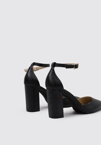 Zapatos de tacón alto negros con un acabado en cuero liso, diseño de punta cuadrada y correa ajustable en el tobillo, con un tacón ancho.