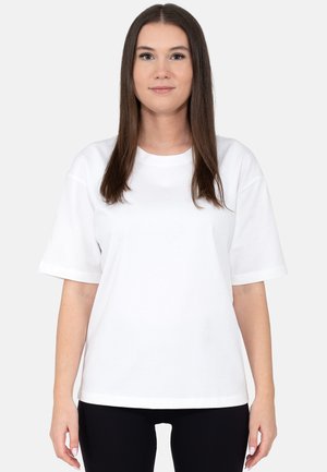 Młoda kobieta z długimi, prostymi włosami, ubrana w prostą białą koszulkę z krótkim rękawem i czarne spodnie, stoi na neutralnym tle.