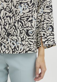 Overhemd met knopen in een zwart en crèmekleurig abstract patroon met lichtblauwe accenten; heeft een rechte snit en zijsplitten op de mouwen.