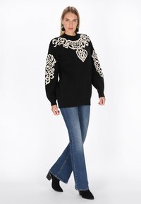 Maglione nero con ricami ornamentali color crema su maniche e scollatura, abbinato a jeans a zampa d'elefante blu e stivali neri con tacco alla caviglia.
