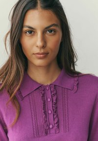 Purple knit polo shirt featuring a textured collar, ruffle accents, and button detailing along the front, highlighting its intricate patterns.