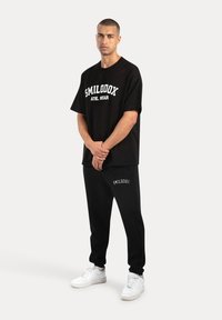 Camiseta de algodón negra con estampado blanco "SMILODOX ATHL. WEAR" y pantalones deportivos negros a juego, con detalle del logo en el dobladillo y zapatillas blancas.