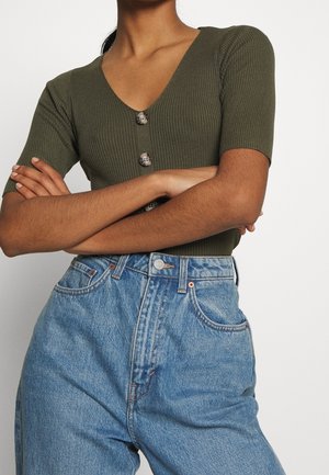 Kobieta w oliwkowo zielonym prążkowanym topie z krótkim rękawem i trzema guzikami, z założonymi skrzyżowanymi rękami, nosząca jasnoniebieskie, wysokowarstwowe dżinsy.