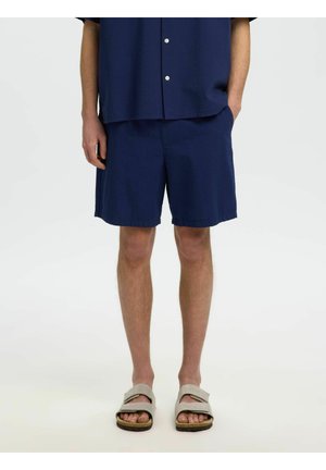 Mężczyzna w granatowych szortach i pasującej koszuli z beżowymi sandałami, stojący na tle jednolitego jasnego tła.