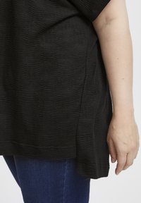 Sort tekstureret top med et bølget mønster, korte ærmer og en løs pasform, parret med mørkeblå jeans i den nederste del af billedet.