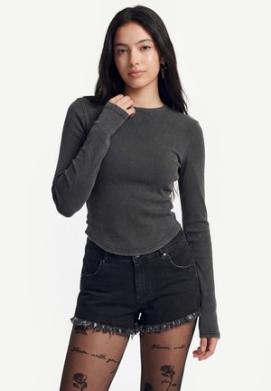 Nuori nainen pitkine tummine hiuksineen, yllään harmaa pitkähiha ja mustat farkkushortsit sekä läpikuultavat sukkahousut, joissa ruusu- ja tekstitatuointeja.