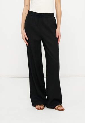 Osoba ubrana w czarne, szerokie spodnie z wiązanym w pasie sznurkiem i brązowe sandały, stojąca na tle jednolitego białego tła.