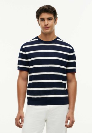 Junger Mann trägt einen kurzärmeligen Pullover mit horizontalen marineblauen und weißen Streifen und weiße Hose, steht vor einem einfarbigen Hintergrund.