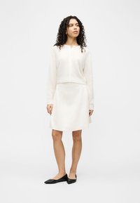 Γυναίκα με σγουρά μαλλιά, φοράει λευκή ζακέτα με κουμπιά και ταιριαστή φούστα, στέκεται με τα χέρια χαλαρά, κοιτάζει αριστερά, φορά μαύρες μοκασίνια.