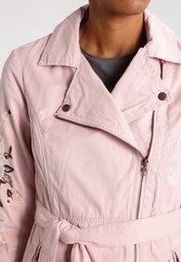 Růžová vzorovaná bunda se zipem vpředu, velkým límcem a páskem v pase. Na levém rukávu má květinovou vyšívku. Hladký povrch.