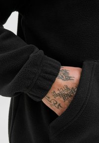Sort fleece pullover med ribbede manchetter og lommer. Glat, blød tekstur med synlig detaljeret syning. Hud med tatoveringer i fokus.