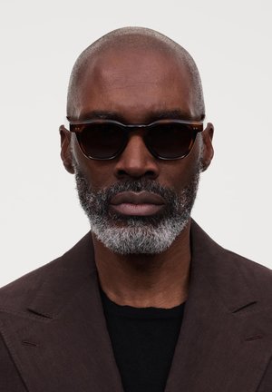 Lunettes de soleil en écaille brune avec des verres ronds, monture sombre et finition mate, portées par un homme à la tête rasée de près et à la barbe grise.