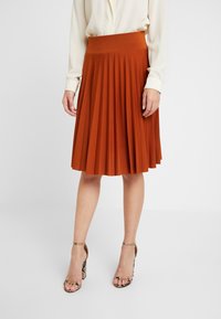 Hnědá plisovaná sukně s širokým pasem, vyrobená z měkké látky. Nosičná s krémovou blůzou a sandály na podpatku s hadím vzorem.