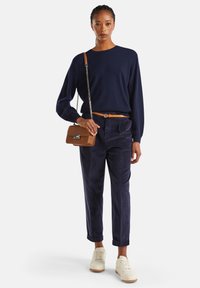 Femme portant un pull marine et un pantalon en velours côtelé, tenant un sac à main marron avec une bandoulière en chaîne, et des baskets blanches, se tenant devant un fond blanc.