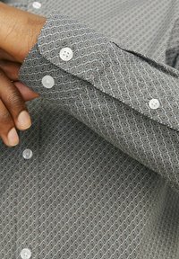 Chemise grise à motif géométrique, avec des boutons blancs et un poignet. La texture du tissu est douce et légère.