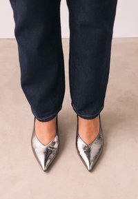 Sötét farmernadrágot és fényes ezüstszínű, hegyes orrú balerinacipőt viselő személy, világos színű padlón állva.