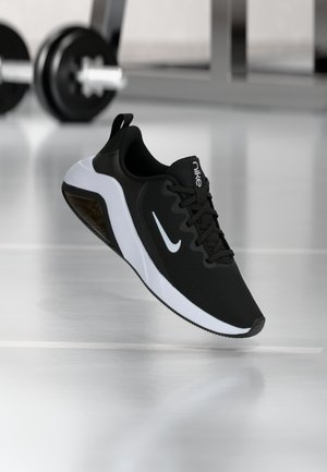 Sort og hvid Nike atletiksko med synlig luftpolster-sål, placeret på en refleksiv gymnastikgulv med slørede vægte i baggrunden.