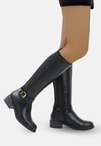 Botas negras hasta la rodilla hechas de cuero suave con un panel texturizado, cremallera lateral y hebilla de acento dorado, con un bajo tacón cuadrado.
