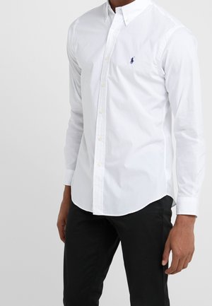 Homme portant une chemise blanche à manches longues avec un petit logo brodé sur la poitrine, assortie à un pantalon noir, debout devant un fond gris.
