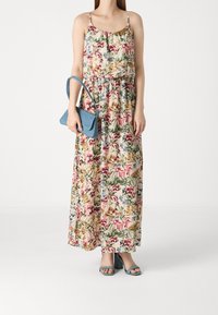 Robe maxi fleurie avec des bretelles fines, présentant un mélange de teintes roses, vertes et dorées. Accompagnée d'un sac à main bleu et de sandales à talons assorties.