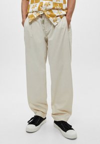 Pantalones de algodón beige de corte relajado, con bolsillos laterales y dobladillos acampanados, combinados con zapatillas negras y una camisa de estampado.