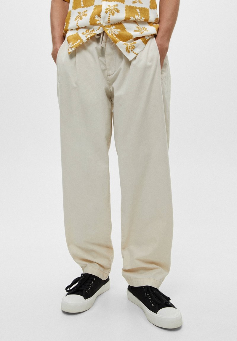 Pantalones de algodón beige de corte relajado, con bolsillos laterales y dobladillos acampanados, combinados con zapatillas negras y una camisa de estampado.