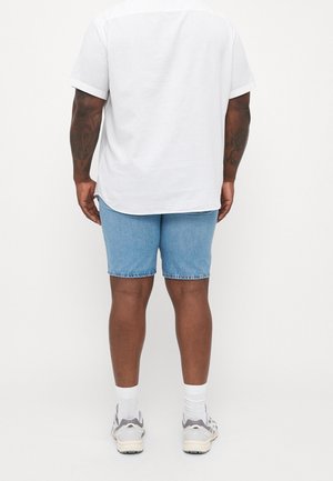 Man wearing a white short-sleeve shirt, light blue denim shorts, white socks, and white sneakers, standing facing away.