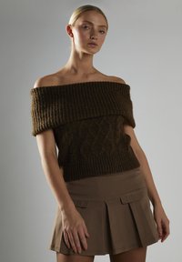 Brązowy sweter z odkrytymi ramionami, dziany, z prążkowanym dekoltem i wzorem w warkocze, zestawiony z jasnobrązową spódnicą plisowaną z kieszeniami.