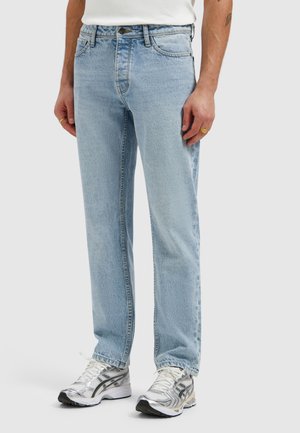 Man die lichtblauwe jeans met rechte pijpen en witte sportschoenen draagt, staand tegen een effen achtergrond.