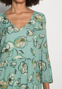 Mujer vistiendo un vestido floral en color verde azulado con escote en V y mangas tres cuartos, frente a un fondo blanco liso.