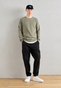 Camisa de manga longa verde-oliva sobre uma t-shirt branca, combinada com calças cargo pretas e ténis brancos, finalizada com um boné preto.