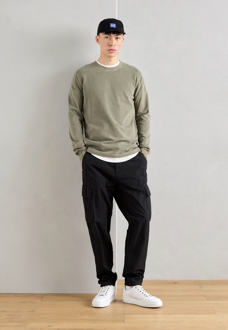 Chemise à manches longues olive superposée à un t-shirt blanc, assortie à un pantalon cargo noir et des baskets blanches, complétée par une casquette noire.