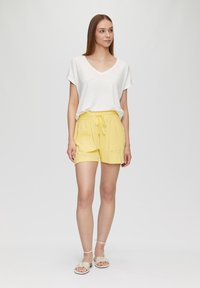 T-shirt blanc à col en V, coupe ample, associé à un short jaune avec cordon de serrage. Le short possède des poches latérales et une texture douce. Des sandales neutres complètent le look.