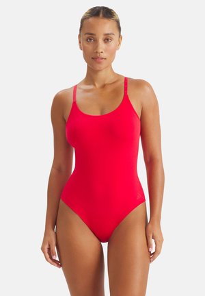 Kvinde med opsat hår iført en skinnende rød badedragt, stående mod en ensfarvet hvid baggrund og kiggende fremad.