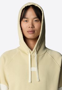 Giovane che indossa un felpa con cappuccio beige e cordini, rivolto in avanti con un'espressione neutra su uno sfondo neutro.