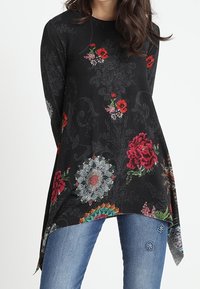 Tunique noire à manches longues ornée de motifs floraux rouges et bleus. L'ourlet est asymétrique. Portée avec un jean bleu décoré d'accents floraux.