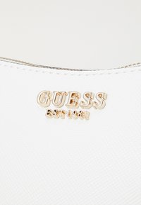 Hvid tekstureret håndtaske med et gyldent "GUESS" logo og detalje "EST 1981". Lynlås lukning øverst med kontrasterende syninger.