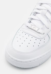 Gros plan sur l'avant d'une basket blanche avec des trous perforés, des lacets blancs et une étiquette en métal sur les lacets portant la mention "AF1".