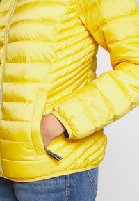 Giacca a vento gialla con finitura lucida, trapuntata orizzontalmente, tasche laterali e polsini a costine. Zip con un tirante a righe.