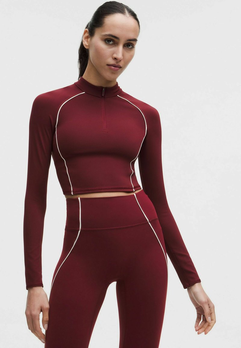 Jeune femme portant un crop top à manches longues ajusté de couleur bordeaux et un legging taille haute avec des détails de coutures blanches, debout devant un fond clair uni.