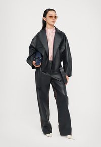 Jachetă oversized din piele ecologică neagră, bluză cu dungi roz, pantaloni largi negri cu accente maronii, pantofi albi cu vârf ascuțit, poșetă bleumarin, ochelari de soare cu lentile colorate.