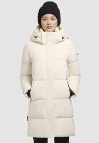 Manteau doudoune long, beige clair, avec un col haut, des poches zippées et une capuche détachable. Présente des panneaux matelassés et des accents noirs.