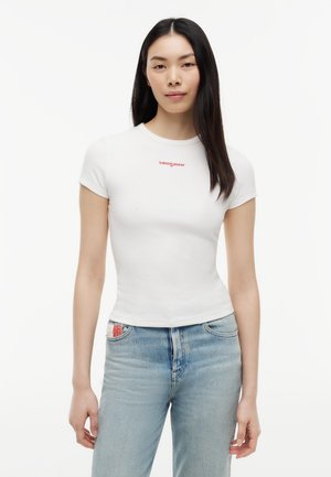 Mujer con cabello largo y negro, vestida con una camiseta blanca ajustada con el logo rojo "tommy jeans" y vaqueros de tiro alto en azul claro sobre un fondo blanco.