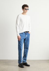 Hvit skjorte med lange ermer, blå jeans, svart belte og svarte sko. Solbriller med rund form. Står mot en enkel bakgrunn.