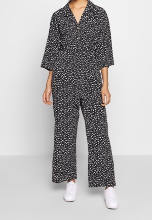 Kvinde iført en sort jumpsuit med hvidt blomsterprint, trekvartlange ærmer, knapper foran, vide ben og hvide sneakers.