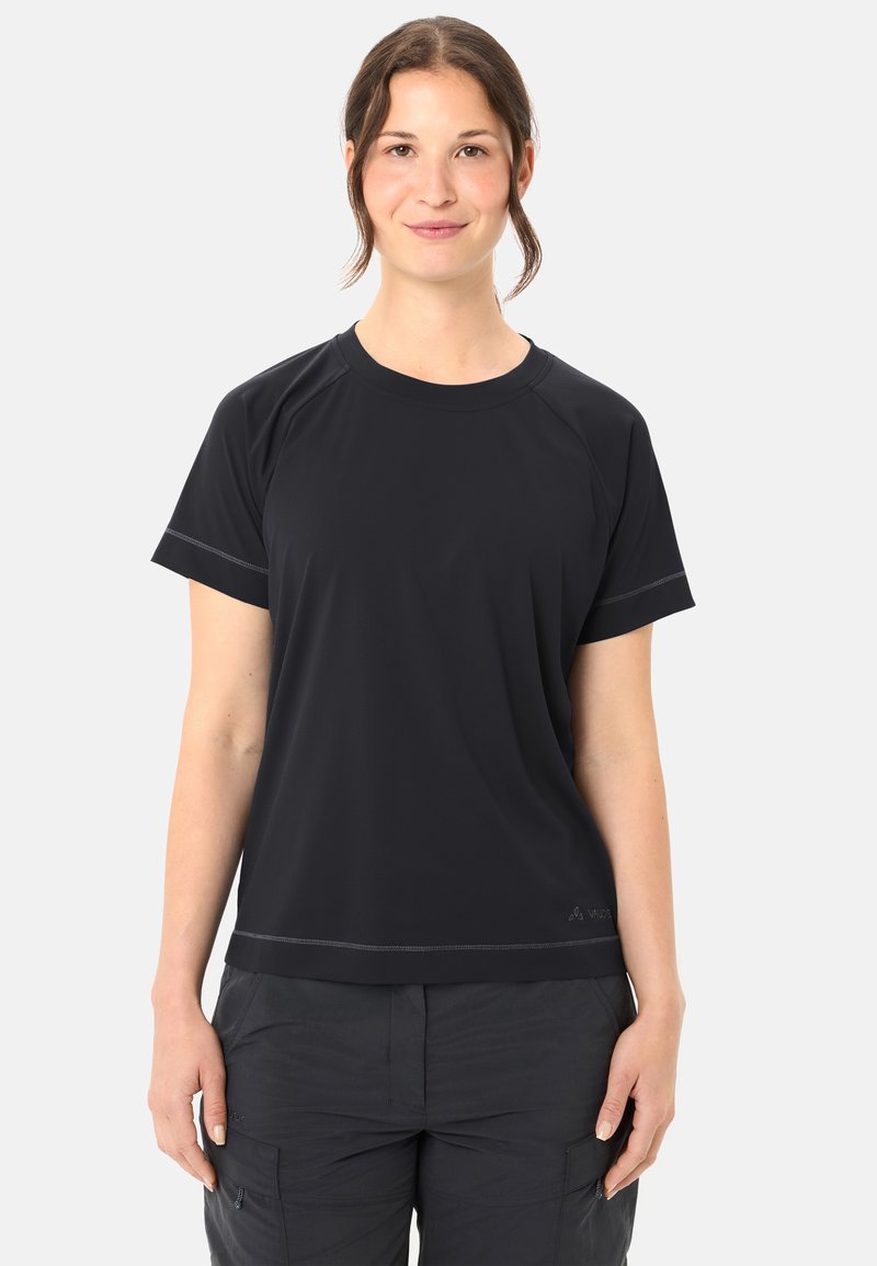 Junge Frau mit braunen Haaren im Pferdeschwanz, trägt ein schwarzes athletisches Kurzarmshirt und dunkle Cargohosen, stehend vor einem schlichten Hintergrund.