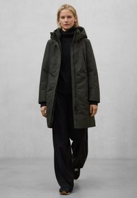 Tmavě zelený prošívaný kabát s kapucí, se zapínáním na přední zip a druky. Nošený přes černý rolák a volné černé kalhoty.