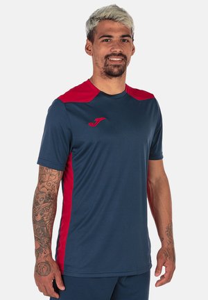 Homme aux cheveux blonds courts et tatouages sur les deux bras portant un maillot de sport bleu marine et rouge, debout et souriant.