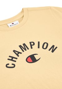 Camiseta de algodón beige con cuello redondo y texto en negro "CHAMPION" con un logotipo en acento rojo. Textura suave y corte casual.